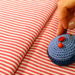 Eine Hand steckt eine Stecknadel in ein Blaues Muffin-Nadelkissen, dass auf einem rot-weiß gestreiften Stoff liegt.