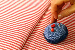 Eine Hand steckt eine Stecknadel in ein Blaues Muffin-Nadelkissen, das auf einem rot-weiß gestreiften Stoff liegt.