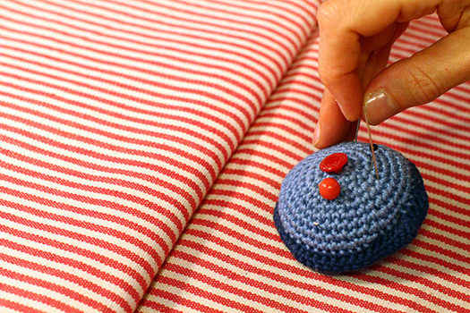 Eine Hand steckt eine Stecknadel in ein Blaues Muffin-Nadelkissen, dass auf einem rot-weiß gestreiften Stoff liegt.